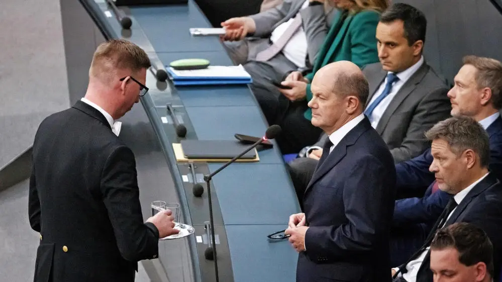 Ein Plenarassistent überreicht ein Glas mit Wasser an Bundeskanzler Olaf Scholz.