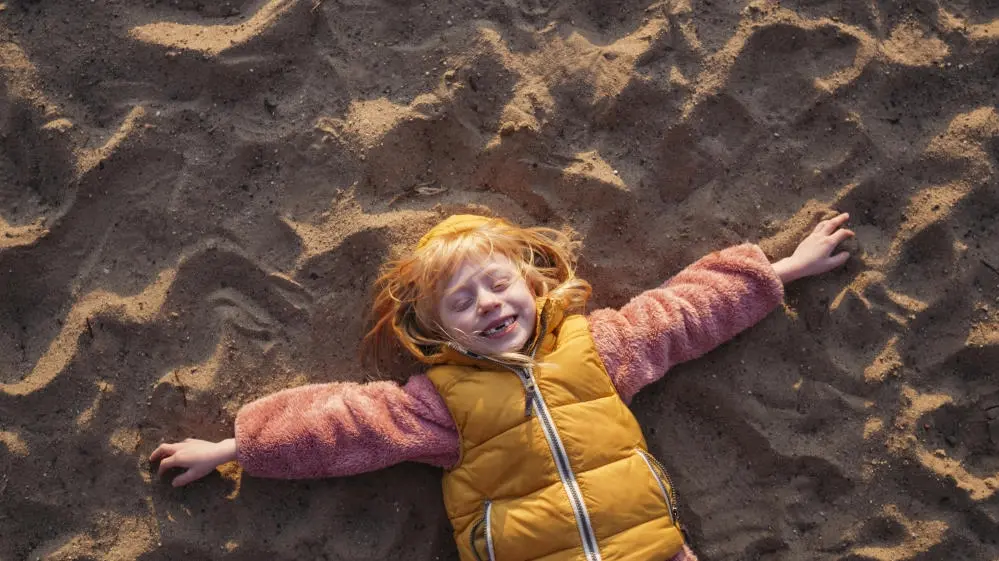 Kind liegt mit ausgestreckten Armen im Sand und lacht