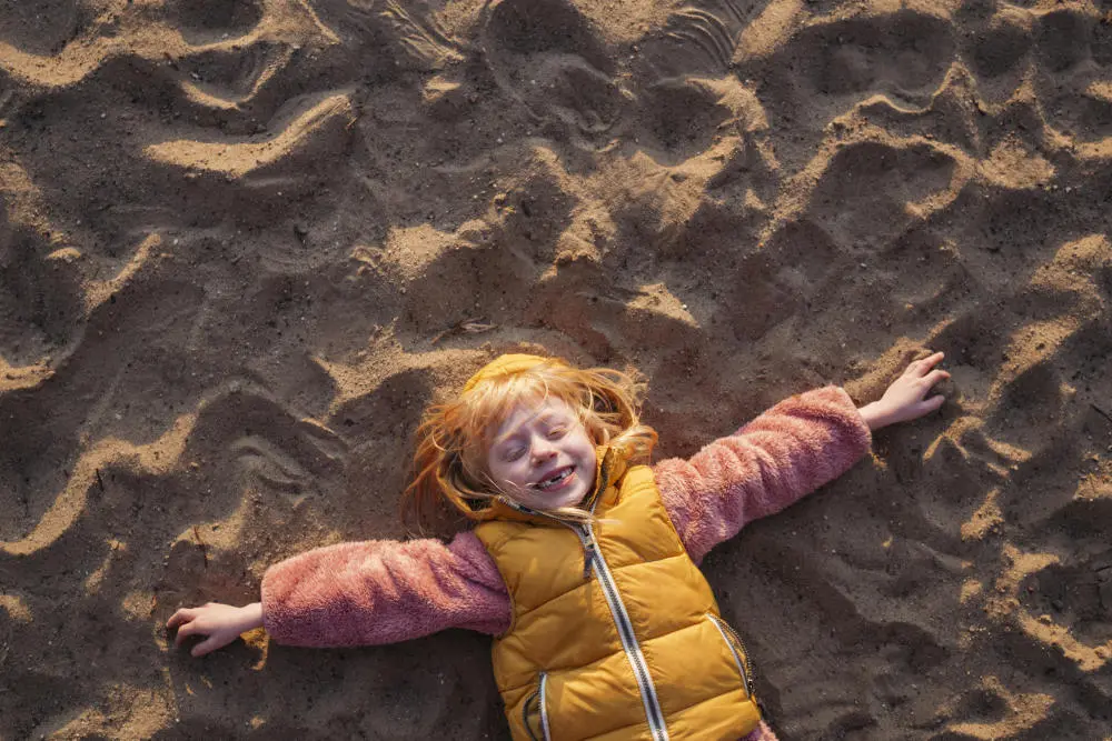 Kind liegt mit ausgestreckten Armen im Sand und lacht