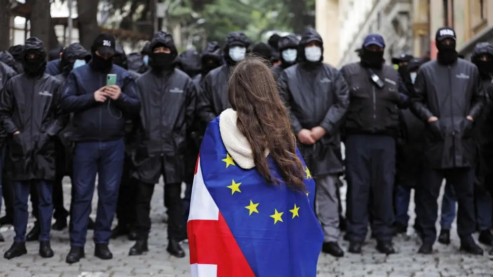 Polizeibeamte stehen während Georgien Protesten Wache. Vor ihnen steht eine Demonstrantin mit umgehängter Flagge.