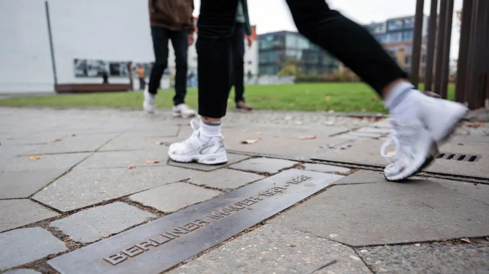 Eine Person geht an der Gedenkstätte Berliner Mauer in der Bernauer Straße über einen Streifen am Boden, der mit "Berliner Mauer" gekennzeichnet ist und den ehemaligen Verlauf nachzeichnet.